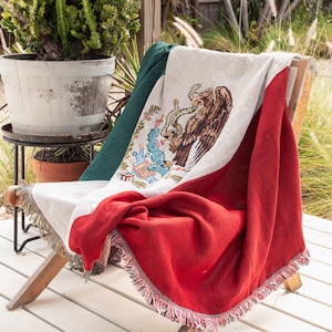 May include: A woven throw blanket featuring the Mexican flag. The blanket is draped over a wooden chair with a fringed edge. The flag is red, white, and green with a brown eagle and cactus design.