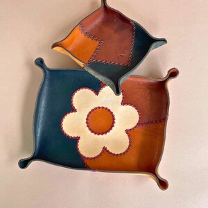 Op de afbeelding: Twee leren dienbladen met een bloemenontwerp. Het grotere dienblad is zwart, bruin en wit met een witte bloem in het midden. Het kleinere dienblad is bruin en oranje. Beide dienbladen hebben rode stiksels.