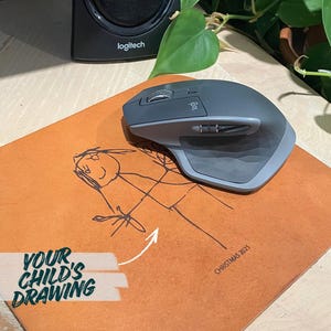 May include: A gray wireless computer mouse rests on an orange mousepad featuring a child's drawing in black ink. The mousepad has the text "YOUR CHILD'S DRAWING" and "CHRISTMAS 2025". A black speaker is in the background.