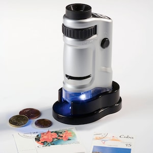 Puede incluir: Un microscopio digital portátil plateado y negro con una luz azul en la base. Varias monedas y sellos postales están al lado del microscopio. Los sellos son de Cuba.