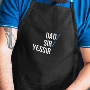 May include: A black apron with white text that reads "DAD/SIR/YESSIR". A person is wearing the apron and holding a knife.