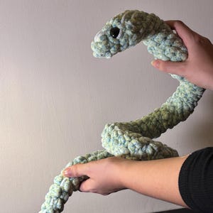 May include: A plush, crocheted snake in shades of blue and green. The snake has a textured, bumpy surface and a black eye. The snake is held up, showing its curved body. The toy is a soft, handmade item.