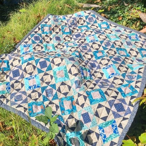 Könnte beinhalten: Eine handgefertigte Steppdecke mit einem geometrischen Muster in Blau-, Türkis- und Beigetönen. Die Steppdecke liegt auf einer grasbewachsenen Fläche in der Nähe eines Gewässers. Das Design zeigt Quadrate, Dreiecke und andere Formen, wodurch ein Patchwork-Effekt entsteht. Der Rand ist ein dunkelblauer Stoff.