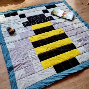 Könnte beinhalten: Eine handgefertigte Steppdecke mit einem Bienenmuster. Die Steppdecke hat einen blauen Rand und ein Patchwork-Design mit schwarzen, gelben und grauen Quadraten. Ein Kinderbuch und ein kleines Spielzeug liegen auf der Steppdecke.