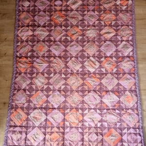 Könnte beinhalten: Eine handgefertigte Steppdecke mit einem geometrischen Muster in Lila-, Rosa- und Pfirsichtönen. Die Steppdecke hat quadratische und rautenförmige Formen mit einem gerüschten Rand. Die Steppdecke besteht aus Stoff mit floralen und unifarbenen Mustern.
