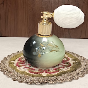 May include: A vintage perfume bottle with a gold atomizer and a white powder puff. The round bottle has a gradient of green and black with gold floral accents. It sits on a decorative, round, multi-coloured doily.