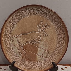Puede incluir: Un plato redondo de cerámica marrón con un diseño en relieve de un ciervo saltando sobre un tronco. El plato tiene un borde decorativo con patrones geométricos. El plato se exhibe sobre un soporte negro.