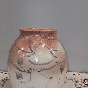 May include: A ceramic vase with a rounded body and a slightly flared rim. The vase is mainly white with a pinkish-brown top and features a hand-painted design of birds and floral elements in blue and brown.