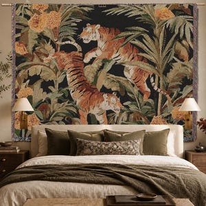 Puede incluir: Un tapiz de pared tejido rectangular que presenta dos tigres en un entorno de selva exuberante con flores naranjas y hojas de palma verdes sobre un fondo negro. El tapiz tiene un borde con flecos y cuelga de una barra met&aacute;lica horizontal.