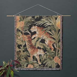 Puede incluir: Un tapiz de pared tejido rectangular que muestra dos tigres en un entorno de selva con hojas de palma verdes y flores naranjas sobre un fondo negro. El tejido tiene un borde con flecos y cuelga de una varilla de madera con un cordel blanco.