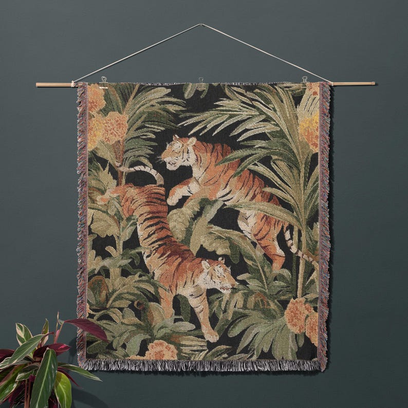 Puede incluir: Un tapiz de pared tejido rectangular que muestra dos tigres en un entorno de selva con hojas de palma verdes y flores naranjas sobre un fondo negro. El tejido tiene un borde con flecos y cuelga de una varilla de madera con un cordel blanco.