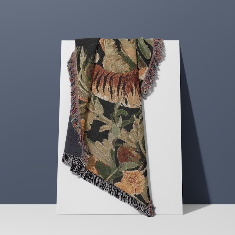 Puede incluir: Una manta tejida con un patr&oacute;n detallado de tigre entre un follaje exuberante en tonos negro, verde, naranja y marr&oacute;n. El tejido rectangular tiene una superficie texturizada y est&aacute; acabado con bordes decorativos de flecos en todos sus lados.