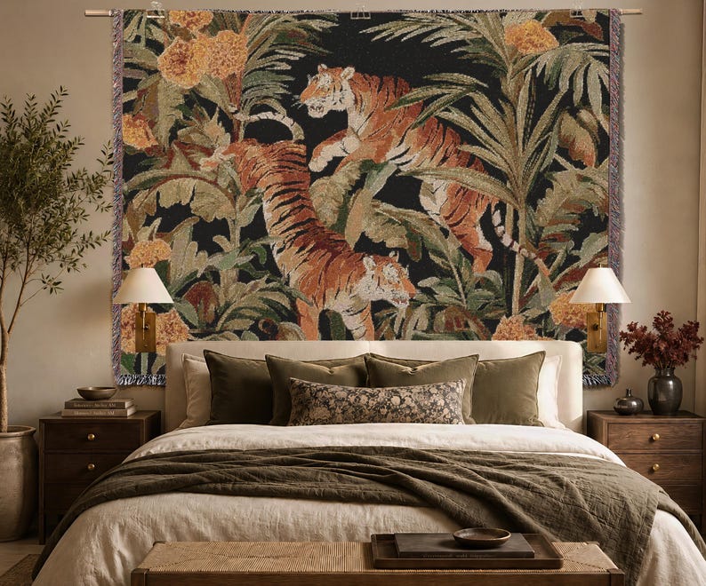 Puede incluir: Un tapiz de pared tejido rectangular que presenta dos tigres en un entorno de selva exuberante con flores naranjas y hojas de palma verdes sobre un fondo negro. El tapiz tiene un borde con flecos y cuelga de una barra met&aacute;lica horizontal.