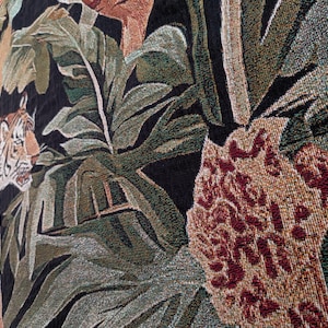Puede incluir: Un tapiz tejido que presenta un tigre con rayas naranjas y negras posicionado entre grandes hojas tropicales verdes y follaje. El dise&ntilde;o incluye un fondo oscuro texturizado y un elemento floral marr&oacute;n rojizo en primer plano.
