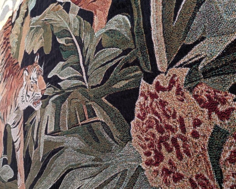 Puede incluir: Un tapiz tejido que presenta un tigre con rayas naranjas y negras posicionado entre grandes hojas tropicales verdes y follaje. El dise&ntilde;o incluye un fondo oscuro texturizado y un elemento floral marr&oacute;n rojizo en primer plano.