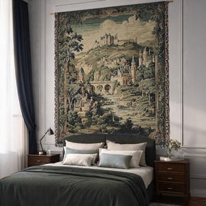 Op de afbeelding: Een groot wandtapijt met een landschap met een kasteel, brug en bomen, hangend boven een bed met een donkergroen dekbed. Het wandtapijt heeft een gedetailleerde, vintage uitstraling met tinten groen, bruin en beige. De slaapkamer heeft donkerhouten nachtkastjes en een zwarte lamp.