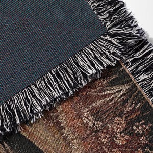 May include: Close-up of a woven throw blanket with a detailed landscape design. The blanket features a dark teal and burgundy textured weave on the reverse side, with a fringe of black and white threads along the edges.