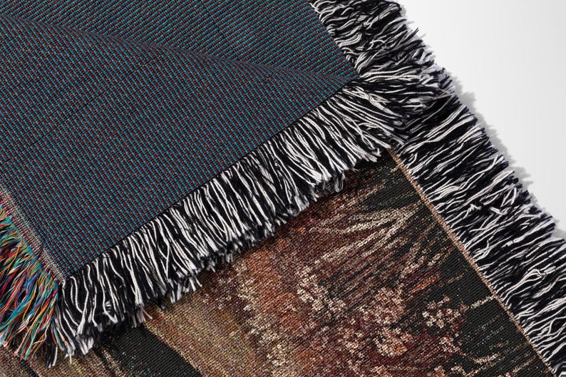 May include: Close-up of a woven throw blanket with a detailed landscape design. The blanket features a dark teal and burgundy textured weave on the reverse side, with a fringe of black and white threads along the edges.