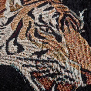 Puede incluir: Un tejido con un dise&ntilde;o detallado de cabeza de tigre. El tigre est&aacute; representado en tonos naranja, blanco y negro, con hilos tejidos visibles que crean la textura. El fondo es negro s&oacute;lido, resaltando el rostro del animal en el tejido.