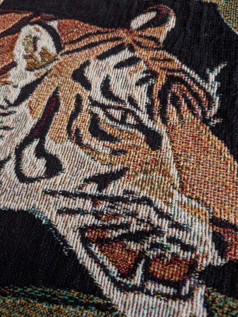 Puede incluir: Un tejido con un dise&ntilde;o detallado de cabeza de tigre. El tigre est&aacute; representado en tonos naranja, blanco y negro, con hilos tejidos visibles que crean la textura. El fondo es negro s&oacute;lido, resaltando el rostro del animal en el tejido.