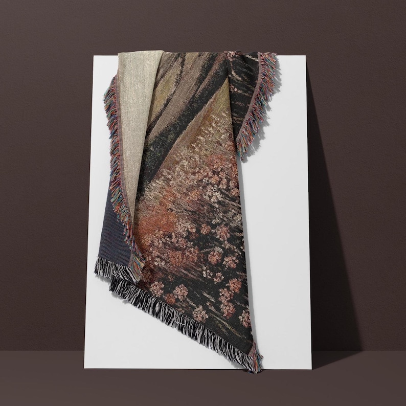 May include: A folded throw blanket with a landscape design. The blanket features a mix of colours, including beige, brown, black, and red, with a fringed edge. The design includes floral and abstract patterns.