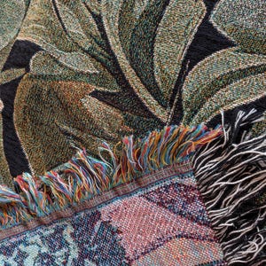 Puede incluir: Una manta textil tejida con un patr&oacute;n bot&aacute;nico de hojas grandes en tonos verde oscuro, oliva y negro. El borde de la tela muestra un fleco decorativo compuesto por hilos multicolores en azul, rojo, amarillo y blanco.