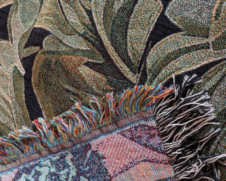 Puede incluir: Una manta textil tejida con un patr&oacute;n bot&aacute;nico de hojas grandes en tonos verde oscuro, oliva y negro. El borde de la tela muestra un fleco decorativo compuesto por hilos multicolores en azul, rojo, amarillo y blanco.