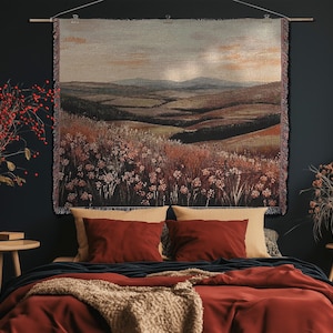 May include: A woven tapestry showing a rural landscape, suspended above a bed with red bedding and cushions. The room is decorated with a vase of red berries and a small wooden table.