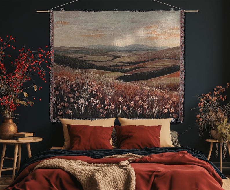 May include: A woven tapestry showing a rural landscape, suspended above a bed with red bedding and cushions. The room is decorated with a vase of red berries and a small wooden table.
