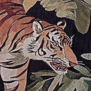 Puede incluir: Un tejido textil con un patr&oacute;n detallado de tigre en tonos naranja, negro y blanco. El tigre aparece representado entre grandes hojas verdes texturizadas sobre un fondo negro s&oacute;lido, mostrando un complejo tejido de hilos multicolor.