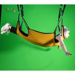 May include: A person suspended in a yellow fabric hammock with black straps. The hammock is hanging from the ceiling against a green background.