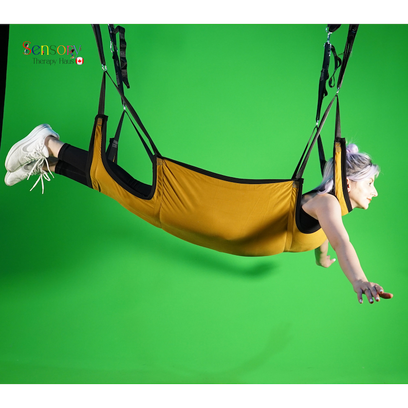 May include: A person suspended in a yellow fabric hammock with black straps. The hammock is hanging from the ceiling against a green background.