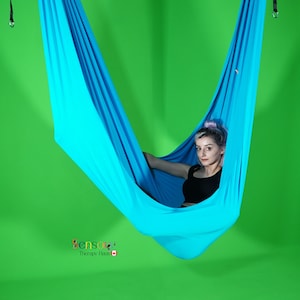 May include: A person sits in a turquoise fabric hammock swing, suspended from the ceiling. The hammock is made of soft, stretchy fabric and has a smooth, even texture. The person is wearing a black tank top and has purple hair. The background is a solid green.