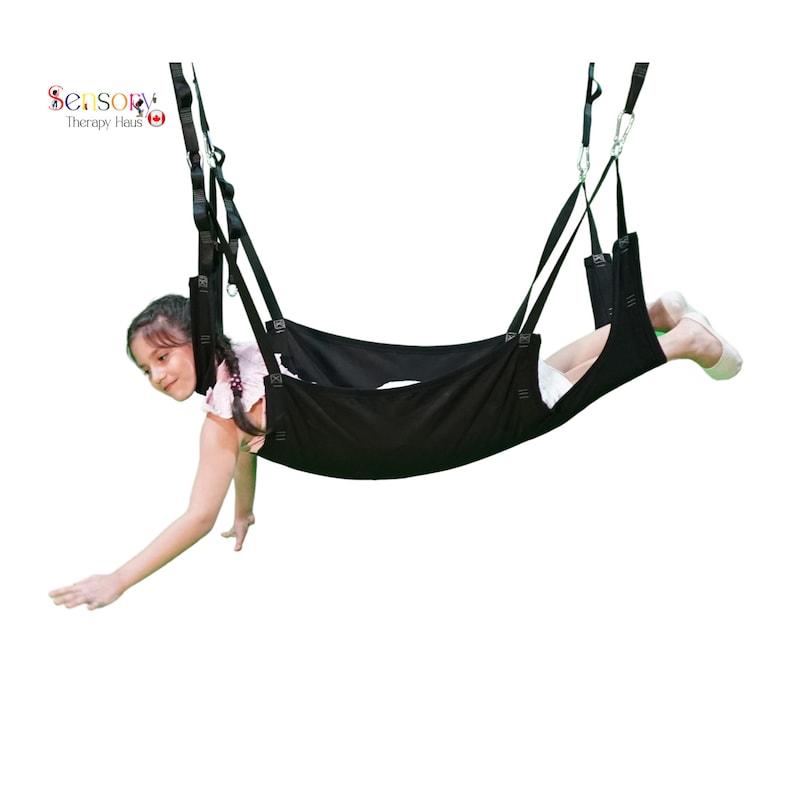 May include: A black sensory swing with a child sitting in it. The swing is suspended from the ceiling by two straps. The child is reaching out with their left hand.
