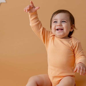 Könnte beinhalten: Ein lächelndes Baby in einem pfirsichfarbenen, gerippten Langarm-Body. Das Baby zeigt mit einem erhobenen Arm nach oben. Der Hintergrund ist einfarbig pfirsichfarben.