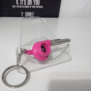 May include: A pink turbocharger key chain with a silver key and a silver key ring. The key chain is resting on a clear plastic stand. The background includes the text "6. IT'S ON YOU" and "7. SMILE".