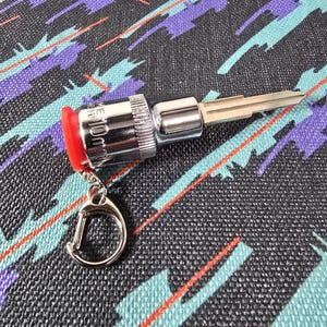 May include: A chrome key with a red rubber cap and a silver keyring. The key is attached to a socket wrench head. The background is a black fabric with a teal, purple, and orange geometric pattern.
