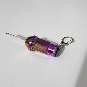May include: A multi-colored D1 Spec lug nut key with a silver key ring. The key has a hexagonal body with a pointed end and a cylindrical end. The key is iridescent, with colors shifting from purple to gold and blue.