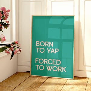 以下が含まれることがあります： 「BORN TO YAP FORCED TO WORK」の文字が入った、額縁に入ったターコイズ色のポスターが木製の床に立っています。 ピンクの花が入ったガラスの花瓶が小さなテーブルの上にあります。 緑の葉が見える窓があります。