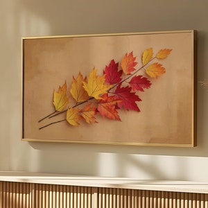 Könnte beinhalten: Ein gerahmter Druck eines Zweigs mit Herbstblättern in Rot-, Orange- und Gelbtönen vor einem beigefarbenen Hintergrund. Der Druck hängt an einer Wand über einem hellen Holzkasten mit einer Lattenfront.