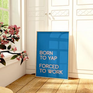 May include: A framed blue poster with the text "BORN TO YAP FORCED TO WORK" stands on a wooden floor. A glass vase with pink flowers sits on a small table near a window. White cabinets are in the background.