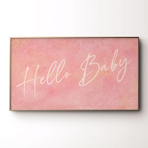 May include: A framed rectangular artwork with a pink watercolor background. The words "Hello Baby" are written in elegant white script across the center. The frame is a thin, gold color.
