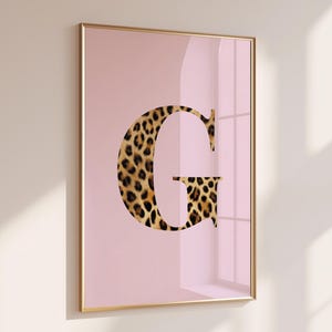 Könnte beinhalten: Gerahmter Kunstdruck mit dem Buchstaben "G" in Leopardenmuster auf weichem rosa Hintergrund. Das Kunstwerk ist in einem goldfarbenen Rahmen eingefasst, was dem Stück Eleganz verleiht. Dieser dekorative Druck eignet sich für die Inneneinrichtung.