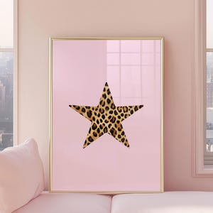 May include: A framed print with a gold border, showcasing a leopard print star on a pale pink backdrop. The artwork is displayed in a room with a window. The star is the focal point, with its brown and black spots contrasting the pink.