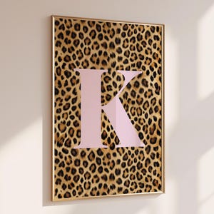 May include: A framed art print featuring a leopard print background with a large, light pink letter "K" in the center. The frame is gold-colored, and the artwork is displayed on a white wall.