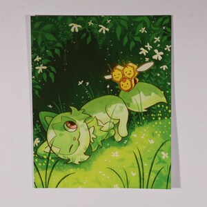 May include: A rectangular art print featuring a green, cat-like creature with large eyes resting in a grassy field. Above the creature, a small, yellow, three-celled honeycomb character with wings and a smiling face hovers in the air.