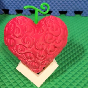 Peut inclure: Un fruit rouge en forme de cœur avec une tige verte et des motifs tourbillonnants. Le fruit est sur un support blanc.