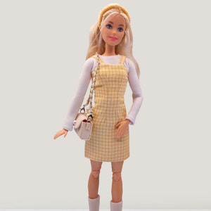 Könnte beinhalten: Eine blonde Puppe, die ein gelbes und weißes kariertes Kleid mit einem weißen Langarmhemd darunter trägt. Die Puppe trägt ein weißes Stirnband und weiße Stiefel. Die Puppe hält eine weiße Handtasche mit einem silbernen Kettenriemen.
