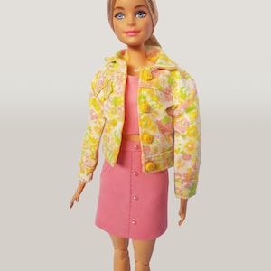May include: A doll dressed in a pink skirt and top, paired with a floral jacket. The jacket features yellow buttons and a white base with yellow and pink floral patterns. The doll has blonde hair and blue eyes.
