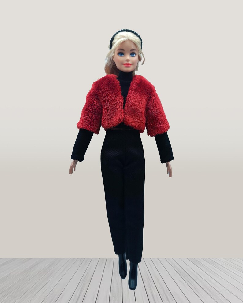 May include: A doll wearing a red faux fur cropped jacket, black turtleneck, and black pants. The doll has blonde hair and is wearing a black headband and black boots. The doll is standing on a light wood floor.